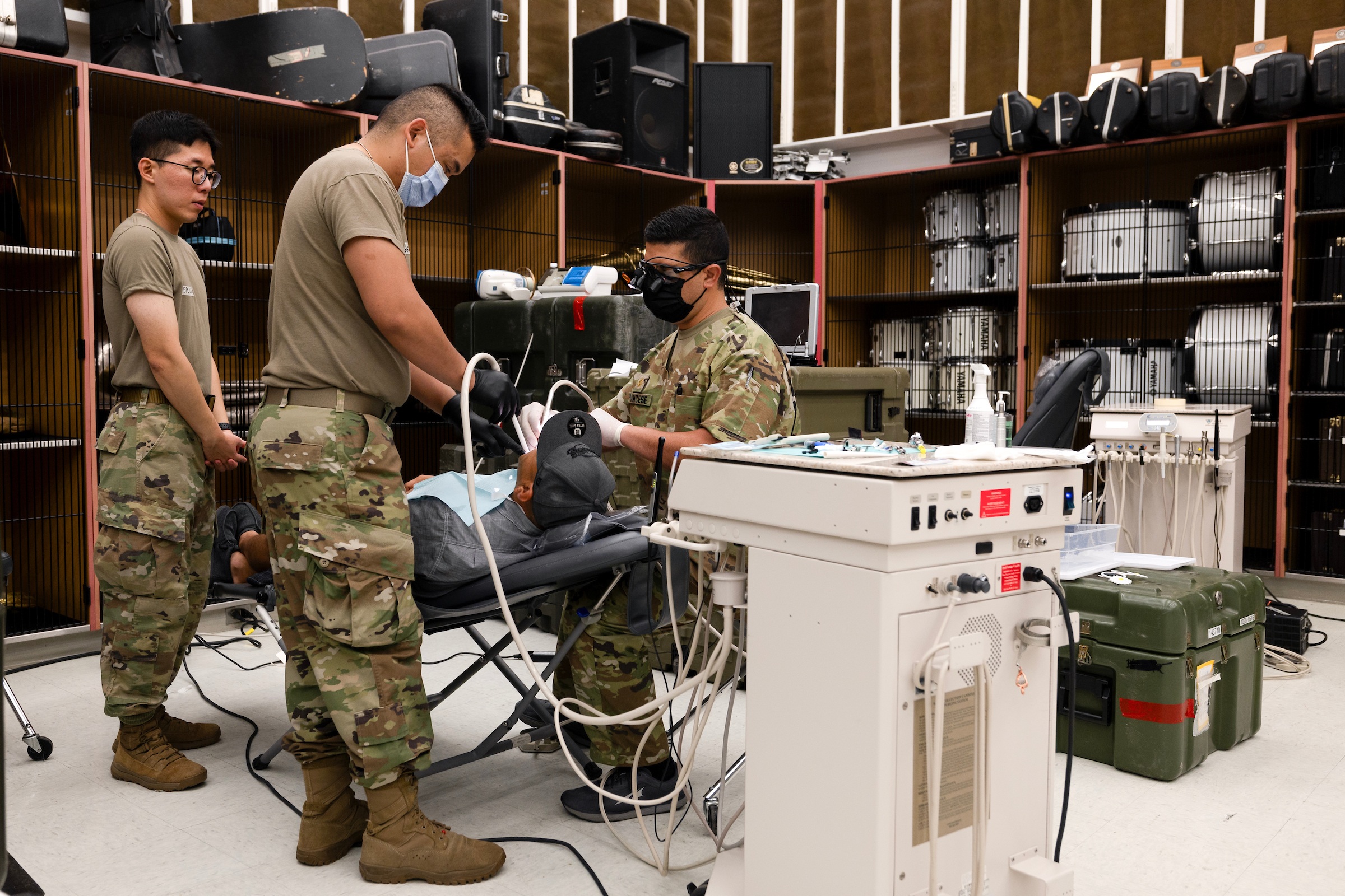 A photo of several people in army fatigues using various dental instruments a reclining man. In the background, drums and other instruments can be seen inside lockers.