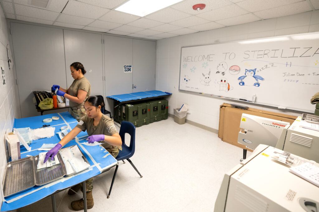 A photo of two young women in army fatigues sanitizing medical instruments. In the background, a large whiteboard reads "WELCOME TO STERILIZATION," with drawings of pokémon and other characters accompanying the text.
