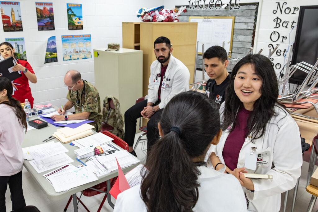 a photo of several students in maroon shirts and lab coats standing around a table talking