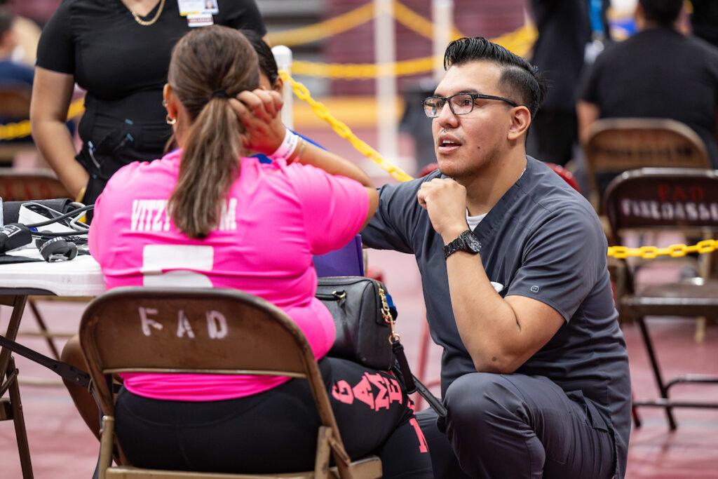 a photo of a man in grey scrubs kneeling down to talk to a woman in a pink jersey