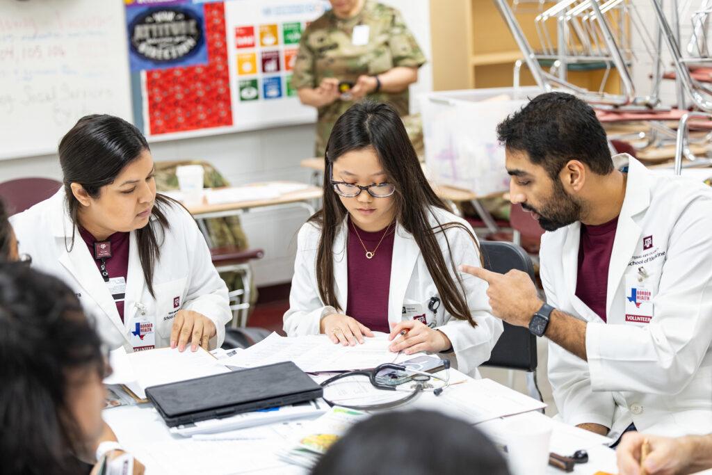 a photo of several students in maroon shirts and lab coats sitting at a table talking