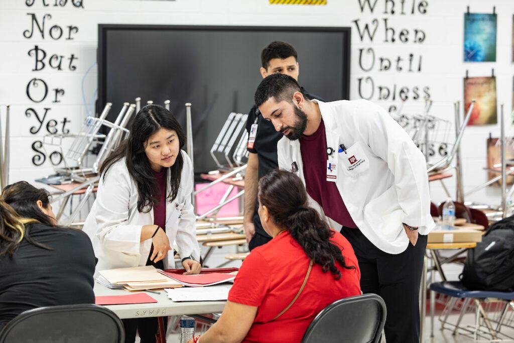 a photo of several students in maroon shirts and lab coats standing around a table talking to a woman in a red shirt