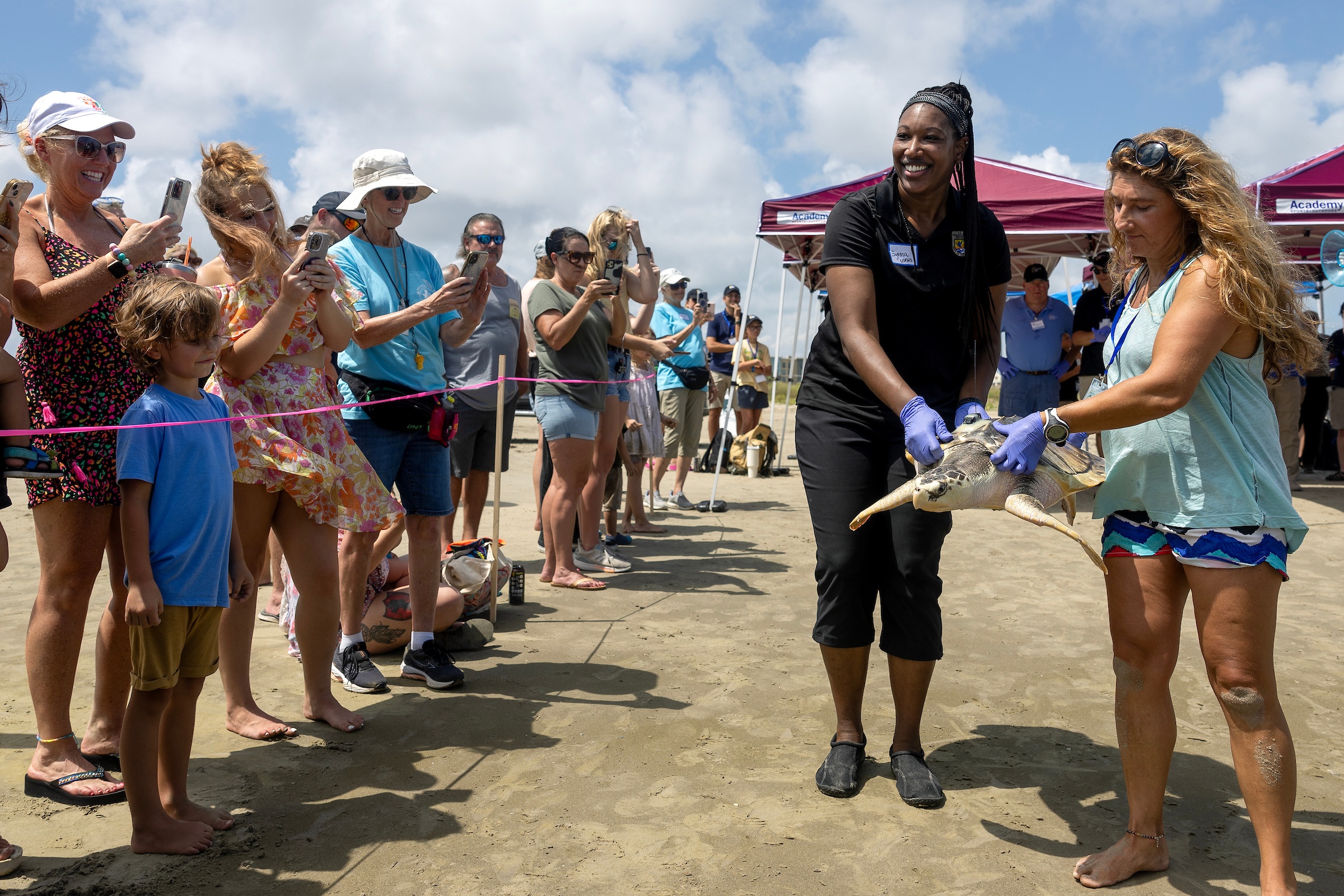 a photo of two woman carrying a sea turtle across a beach as onlookers smile and take photos