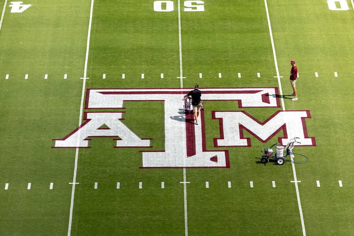 Overhead of Kyle Field at the 50 yard line, where two men paint stripes of maroon paint onto the A&M logo