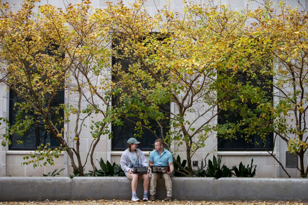 A photo of two people sitting on a concrete ledge under a tree canopy with laptop computers open.
