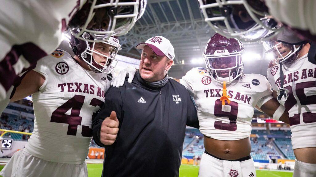 a photo of Mike Elko in Texas A&M gear standing in a huddle with Aggie football players at the Orange Bowl