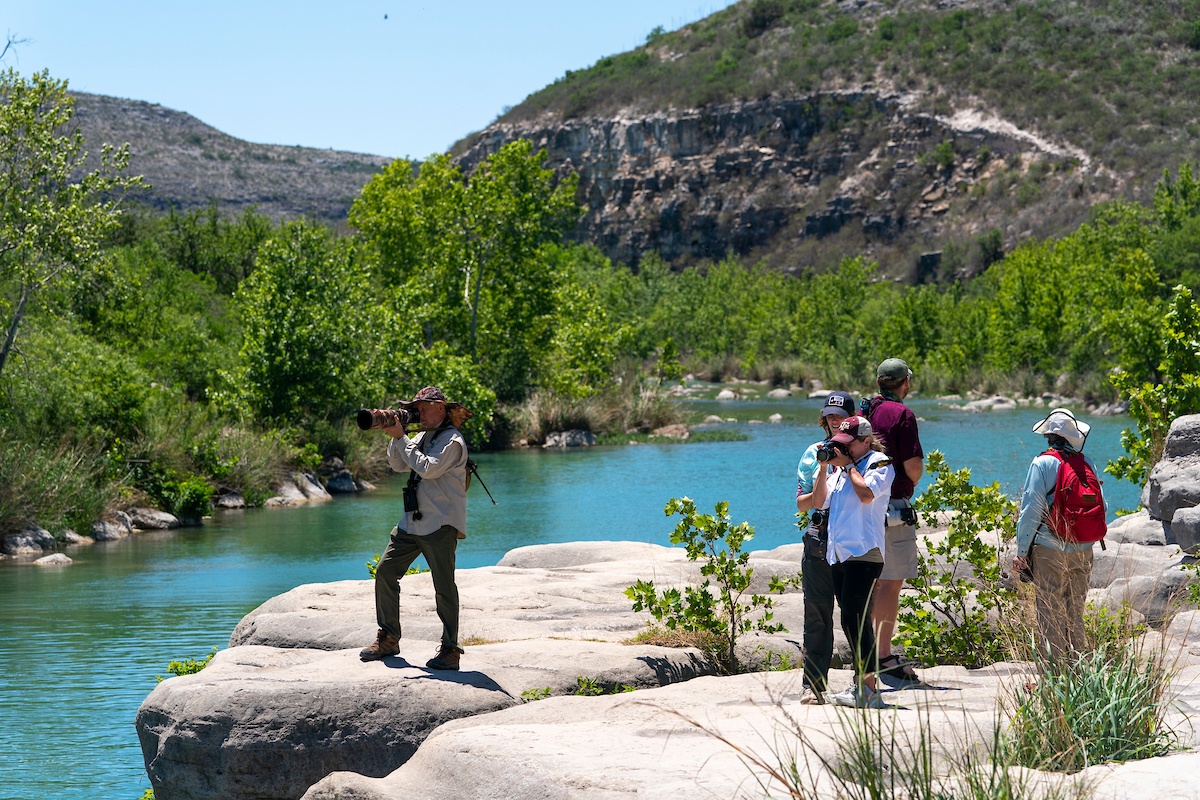 A group of five people take photos and enjoy the scenery along a river in a semi-arid Texas landscape.