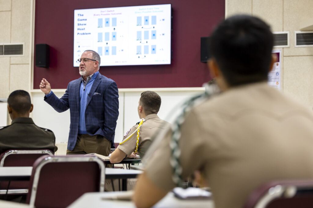 A photo of a man in a classroom with a screen behind him and students in the foreground.