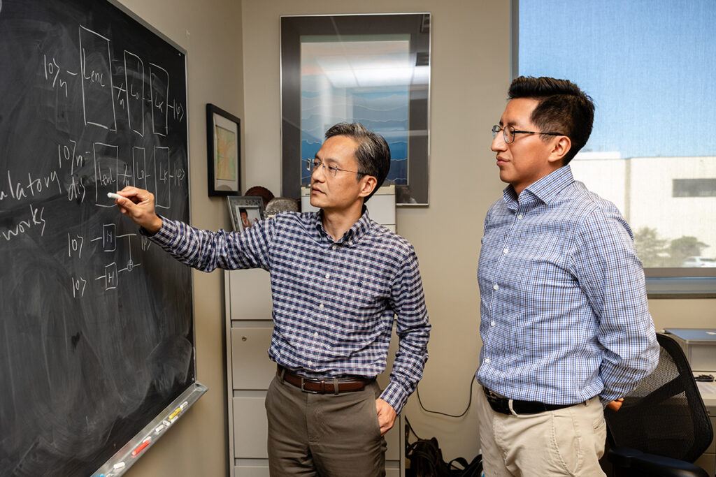 Cai is writing on a chalkboard while Vicharra observes