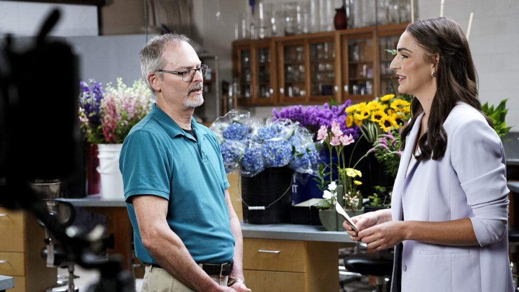a photo of a man and woman talking with a table filled with colorful flowers behind them