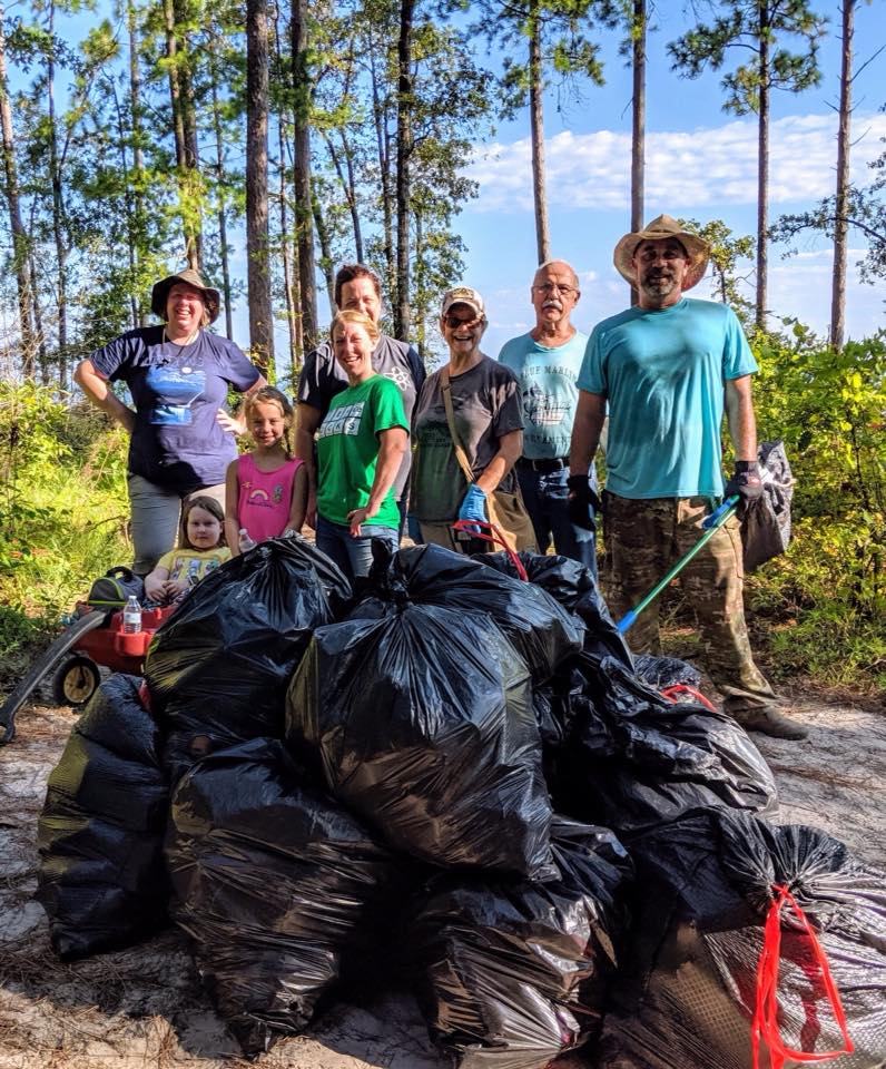 A group of eight volunteers stand behind at least nine bags of litter they collected from the outdoors.