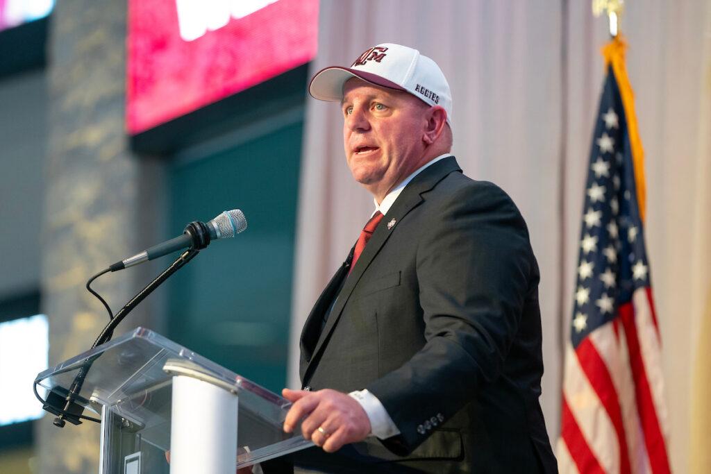 a photo of Mike Elko in a Texas A&M cap speaking at a podium