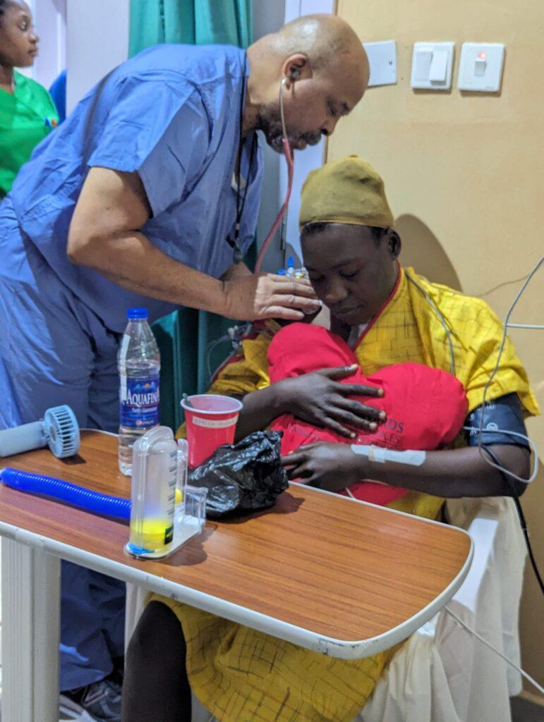 a photo of a man in blue scrubs using a stethoscope to listen a woman's heart while she holds a heart-shaped pillow