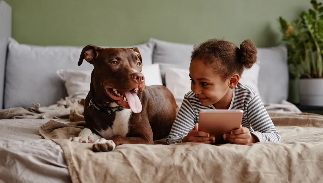 A photo of a young girl in a bed looking at a tablet with a dog next to her on the bed.