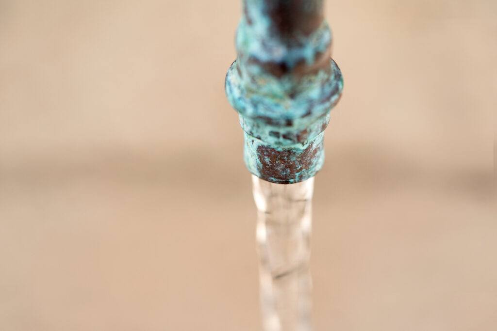 Water flows out off a hose nozzle downward. The nozzle has a patina to it.