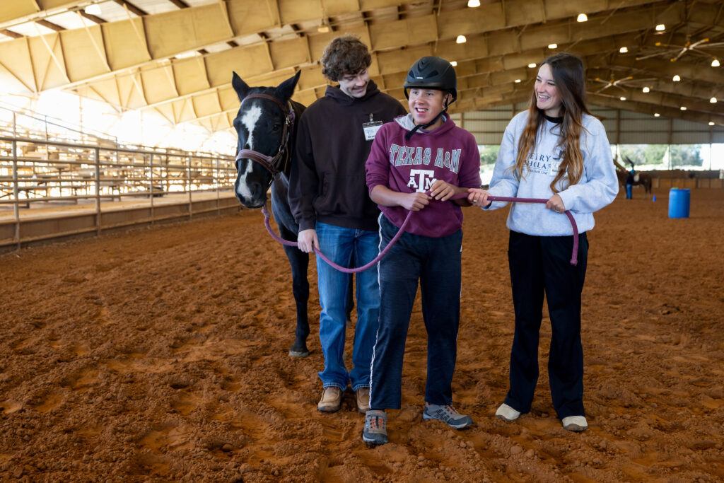 a photo of a young man wearing a helmet and Texas A&M sweatshirt holding a black horse's lead rope alongside two older people