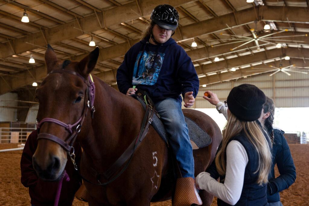 a photo of a young man in a hoodie sitting on a horse and interacting with two women