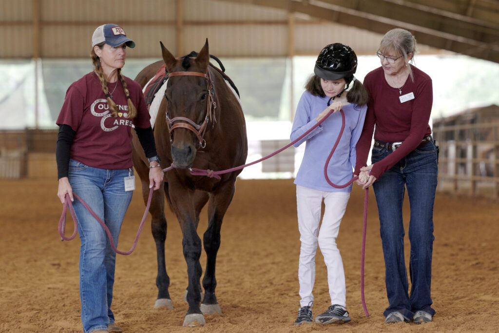 a photo of a woman in a maroon long-sleeve shirt assisting a young girl as she leads a horse across an equestrian arena, as another woman in a cap and Courtney Cares t-shirt looks on