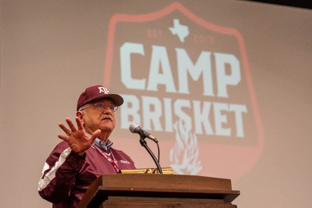 a photo of a man in a maroon Texas A&M cap and jacket speaking at a podium with the Camp Brisket logo behind him