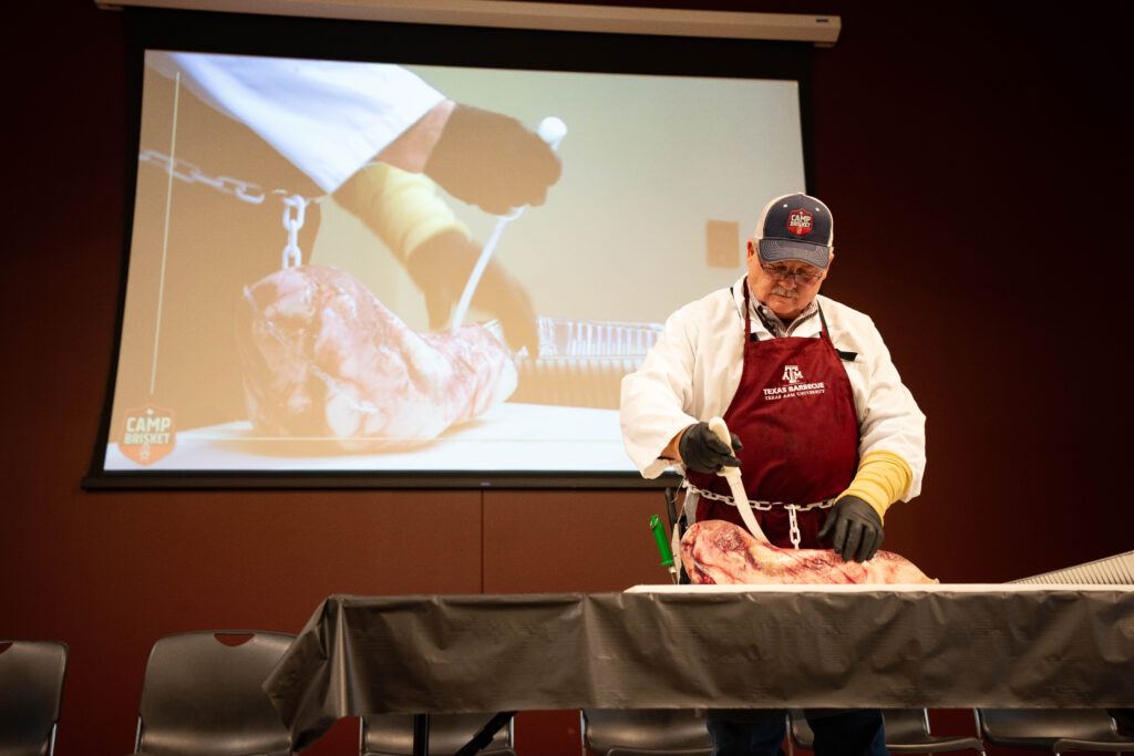 a photo of a man in a maroon Texas A&M apron standing at a table cutting open a packaged brisket