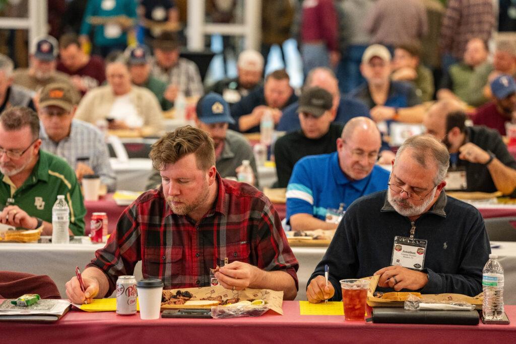 a photo of people sitting behind rows of tables eating brisket and writing