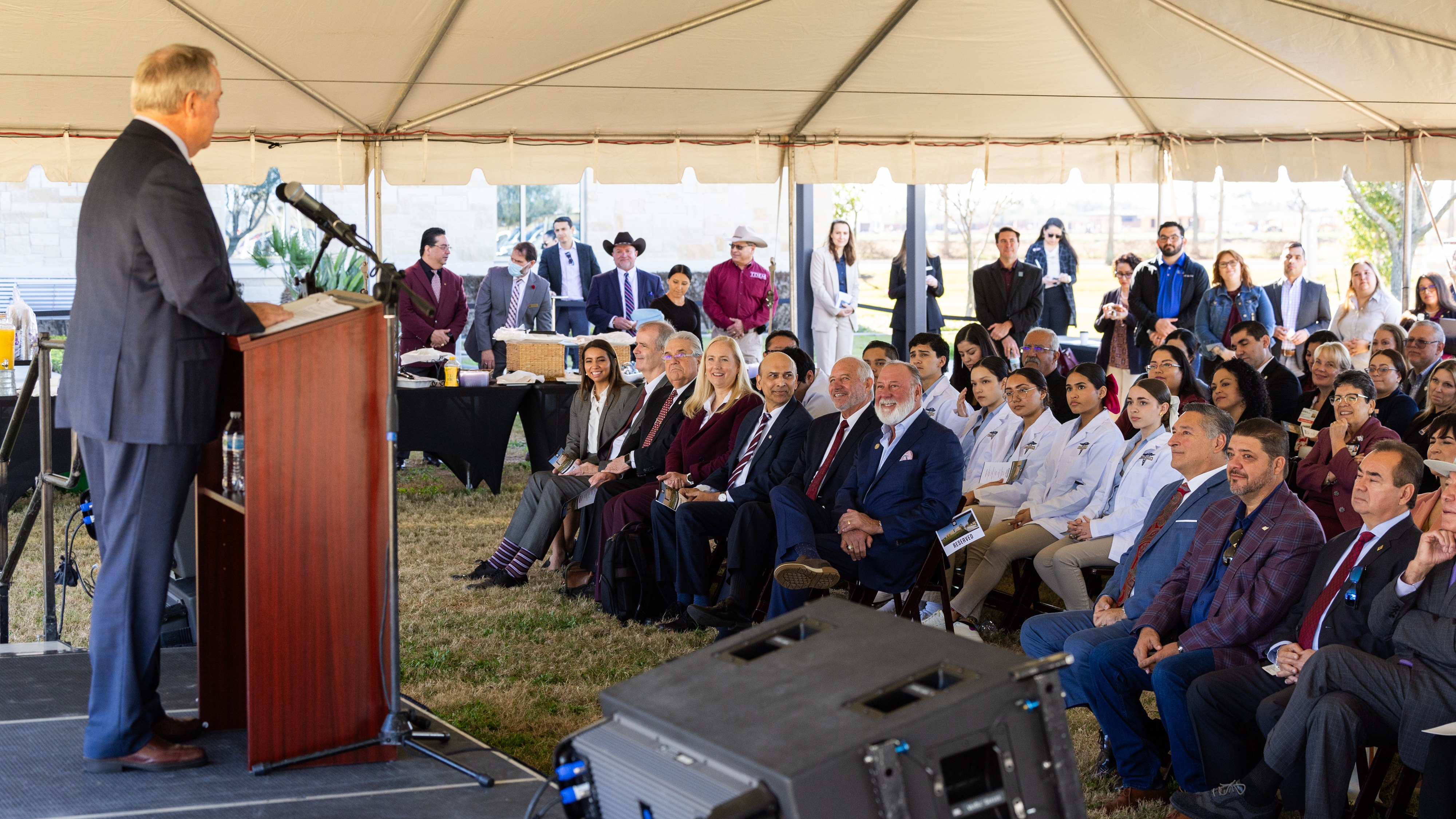 A photo of a speaker at a lectern on a podium speaking to a crowd underneath a tent.