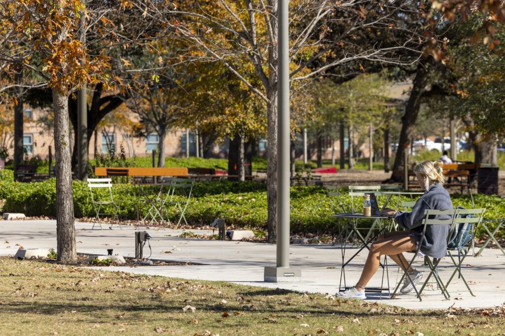 A photo of a woman sitting at a table on a plaza surrounded by trees and fall leaves.