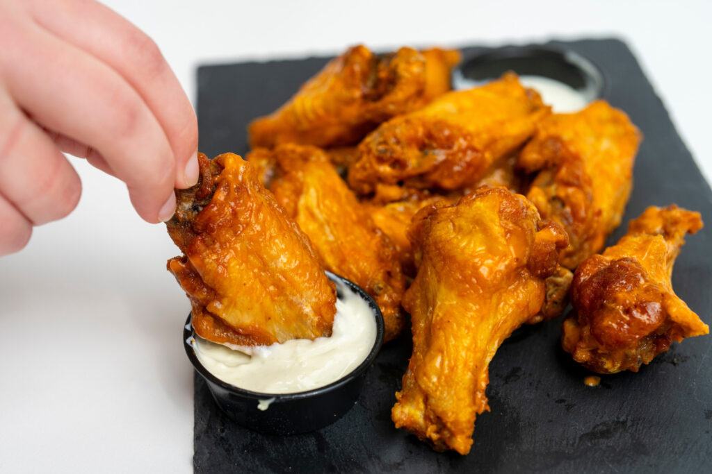 A hand dips a buffalo wing into some dressing with other wings sitting on a plate.