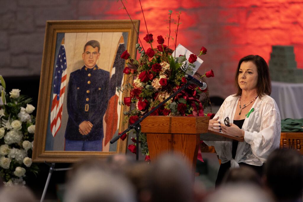A photo of a woman at a lectern speaking at a funeral with a framed painting of a Marine Corps veteran surrounded by flowers to the side of the lectern.