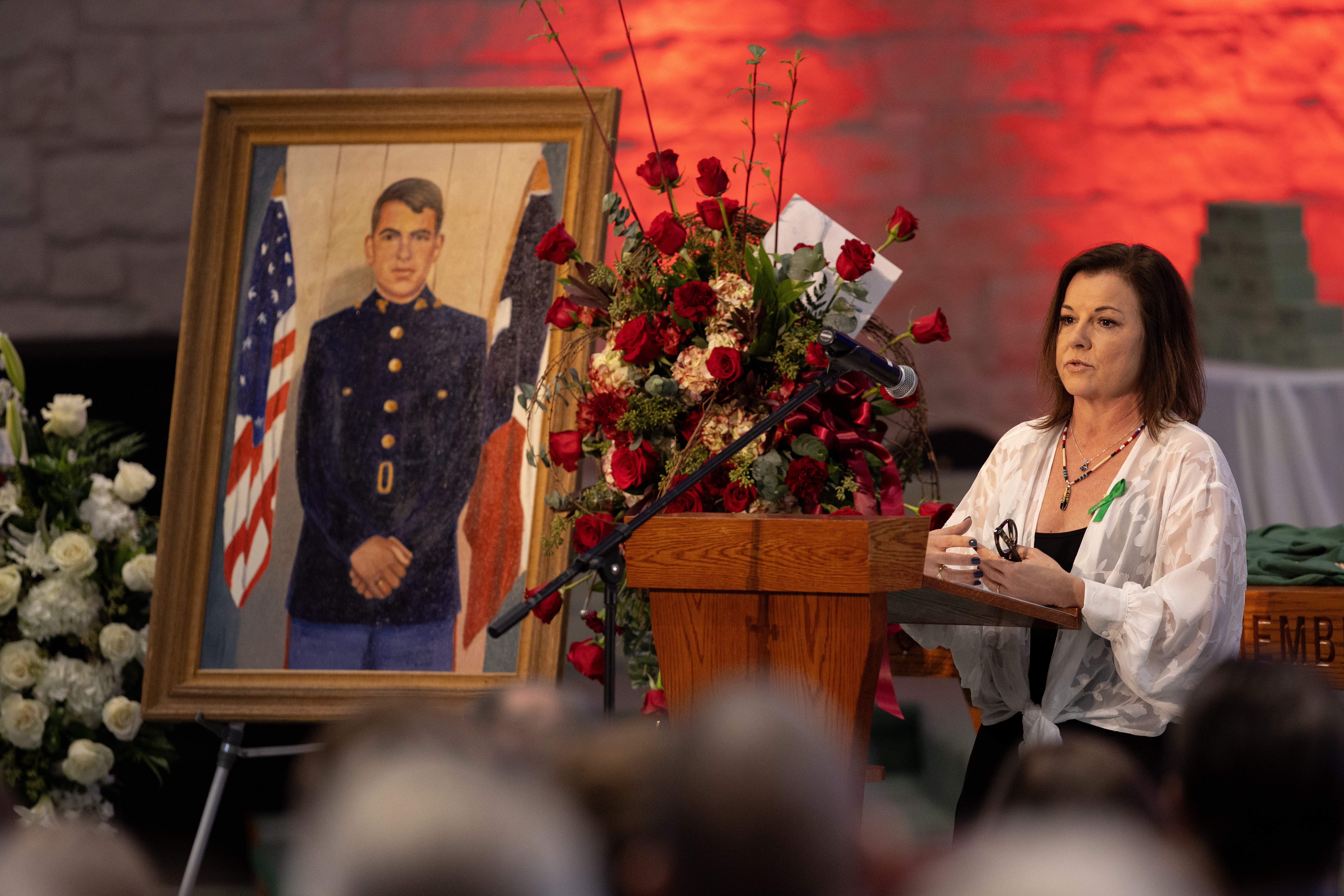 A photo of a woman at a lectern speaking at a funeral with a framed painting of a Marine Corps veteran surrounded by flowers to the side of the lectern.