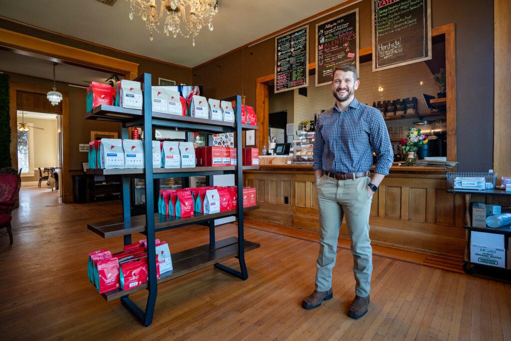 a photo of a man standing next to a shelf full of bags of coffee beans, with several chalk board menus hanging overhead