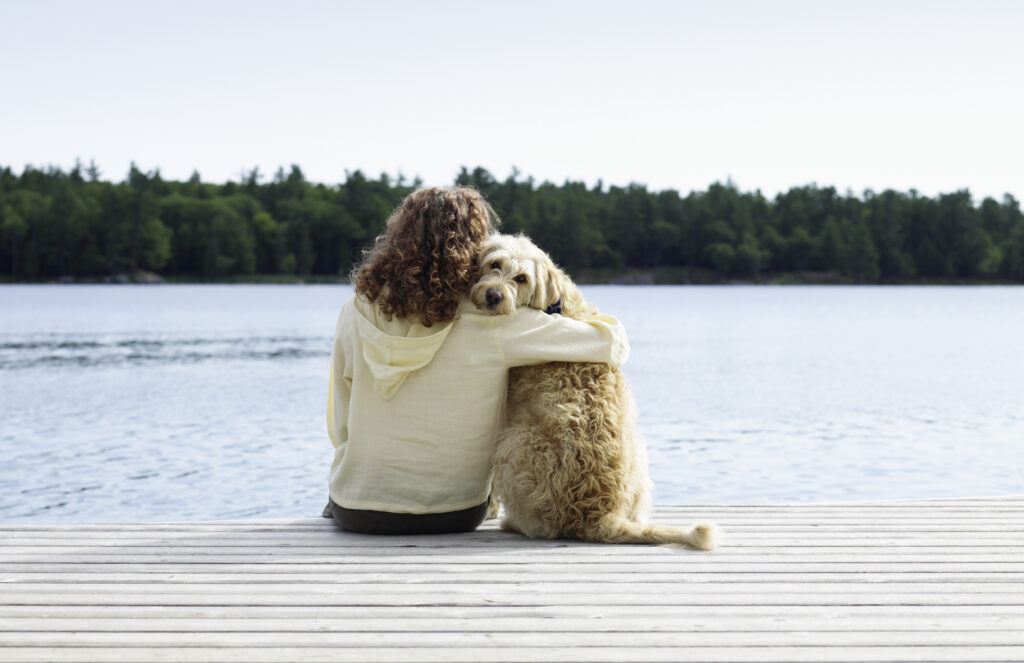 A photo of a person sitting with a dog on the dock of a lake.