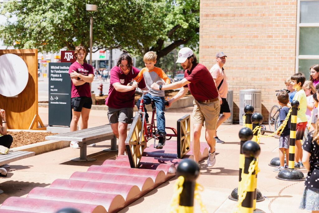 Aggies help a young visitor to ride a bike with square wheels at the 2023 Physics and Engineering Festival