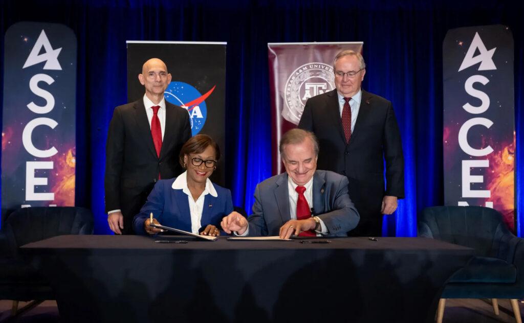 A photo of two people at a table signing papers with two people standing behind them.