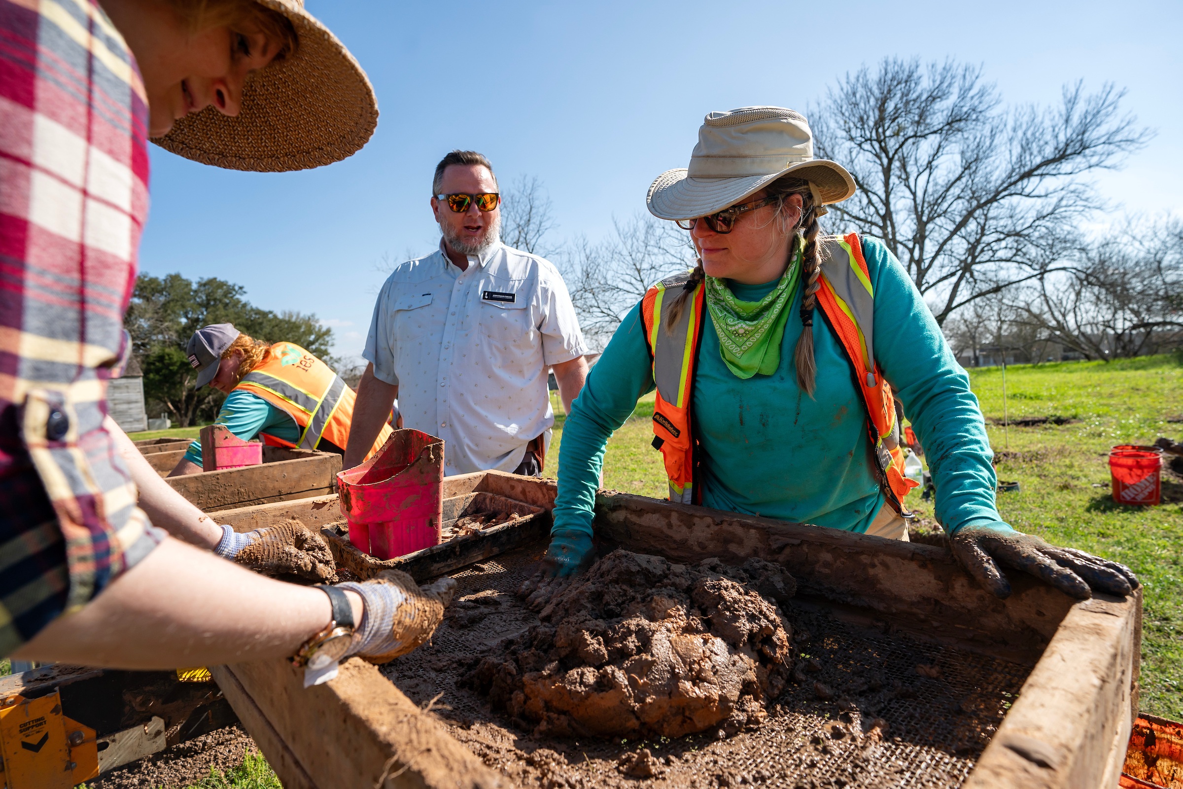 two women in sun hats and garden gloves push mud through a large metal sieve while a man with sunglasses and a name tag speaks with them