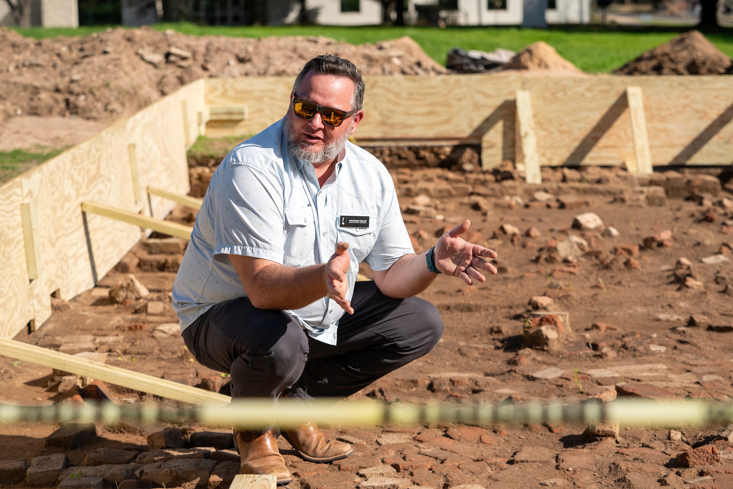 a man in sunglasses and a short sleeved collared shirt crouches on an old brick floor surrounded by plywood