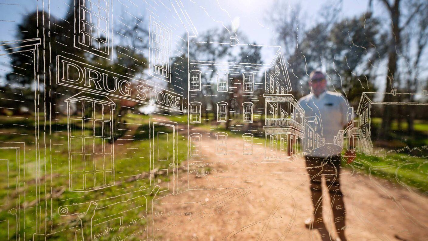 A man stands behind a glass etching of storefronts and other structures with the sun shining overhead