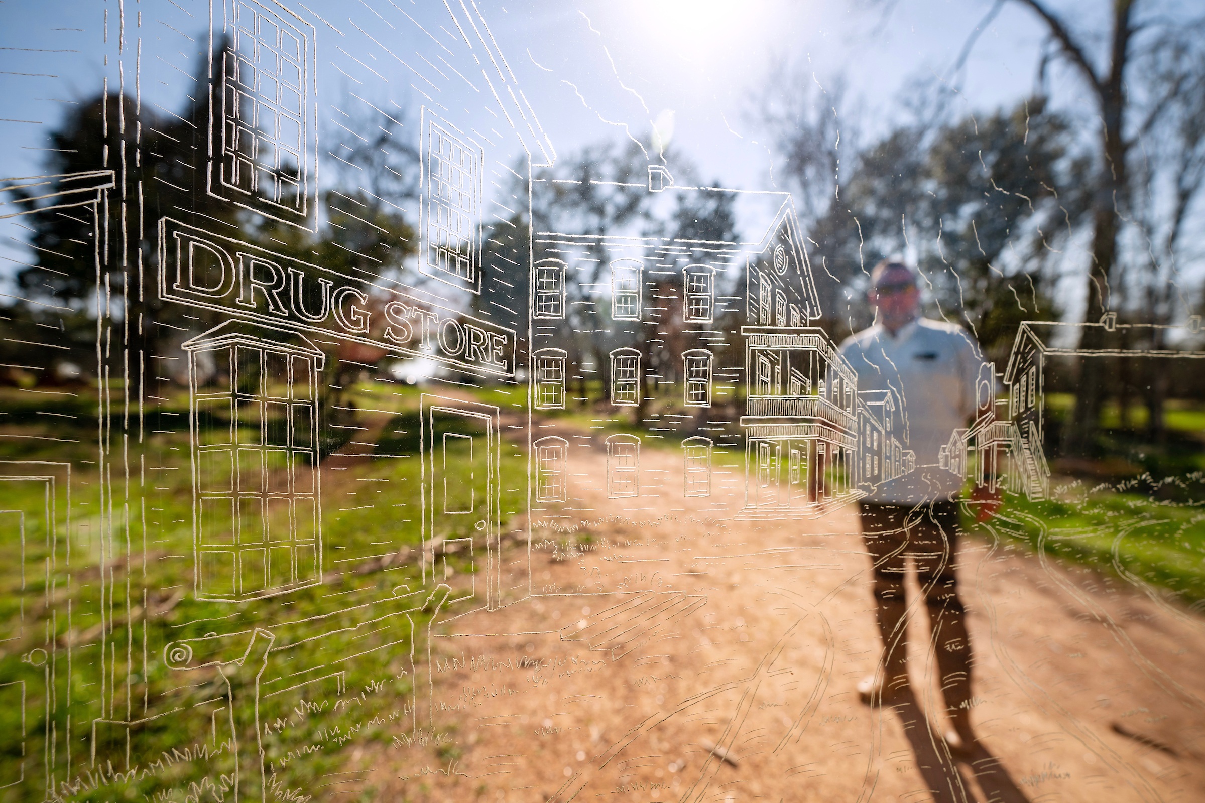 A man stands behind a glass etching of storefronts and other structures with the sun shining overhead