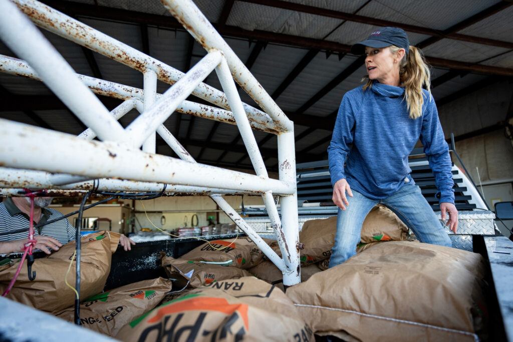 A woman wearing a baseball cap and blue sweatshirt loads bags of feed onto a pallet.