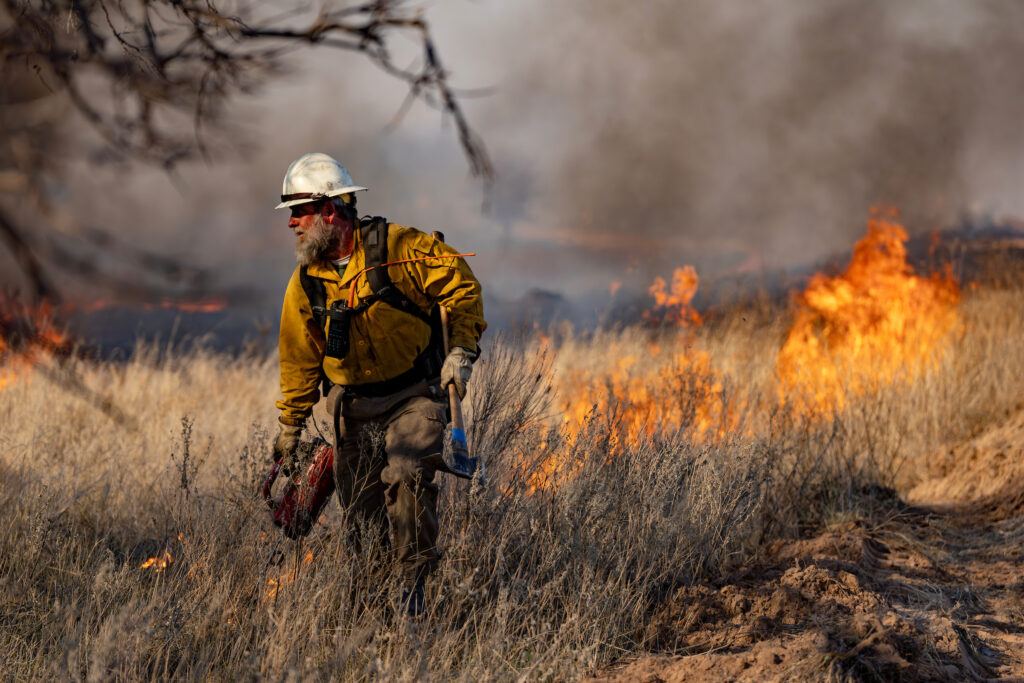 A photo of a man in wildland firefighting gear walking through brush with flames and dark smoke behind him
