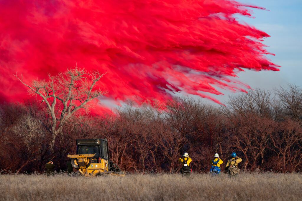 a photo of people in yellow firefighter outfits looking up at a red cloud of fire retardant over a cluster of trees