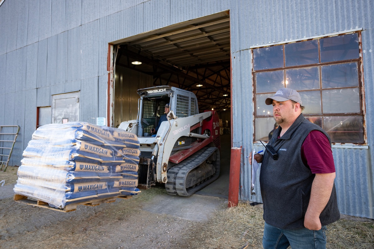 A man stands outside of a building directing a person operating machinery carrying bags of feed out of a supply point.