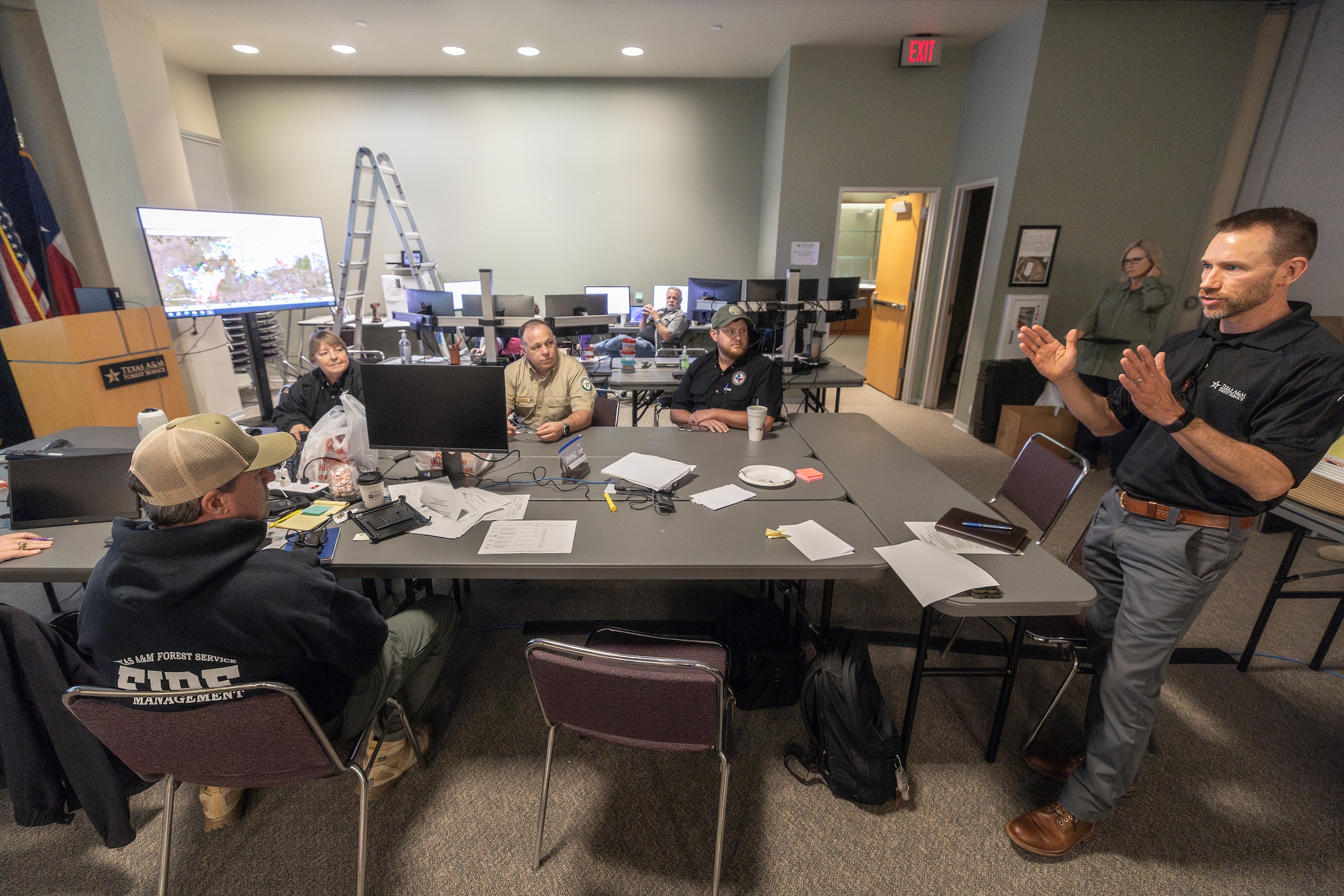 a photo of a man standing at the head of a conference table talking to a small group of people. In the background, a satellite map shows the outline of the Smokehouse Creek fire and other wildfires in the Panhandle.