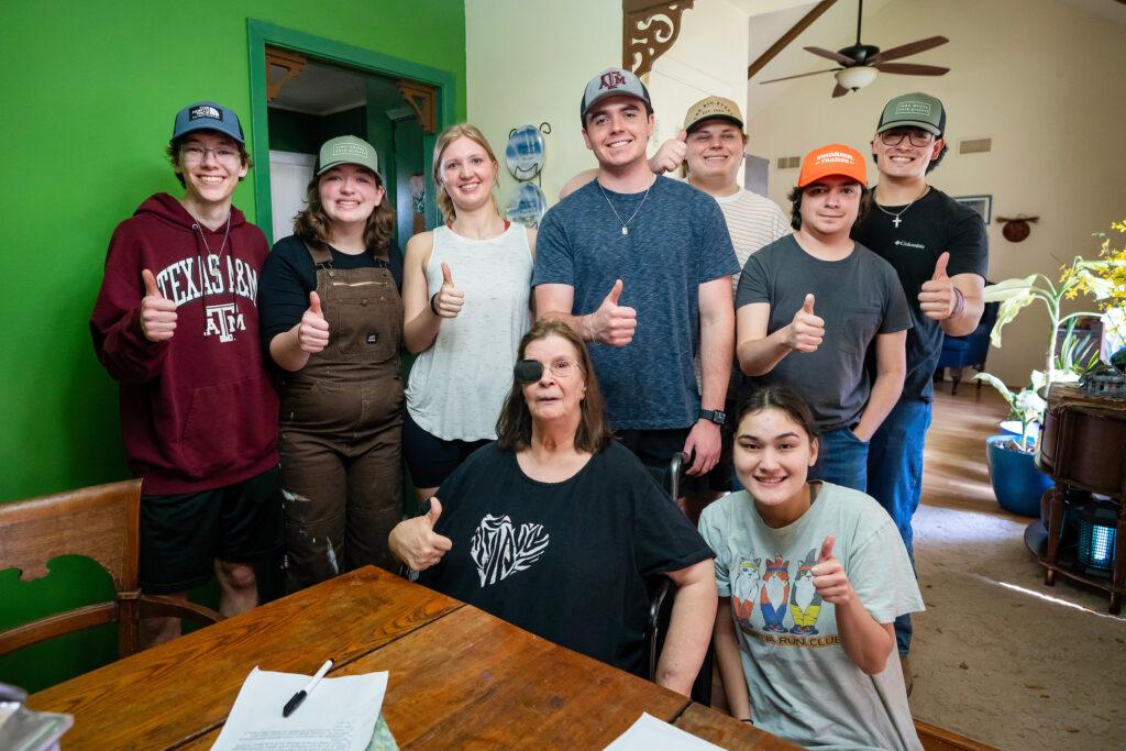 a group of young people stand around an older woman in a wheel chair. all of them are giving the thumbs up and smiling