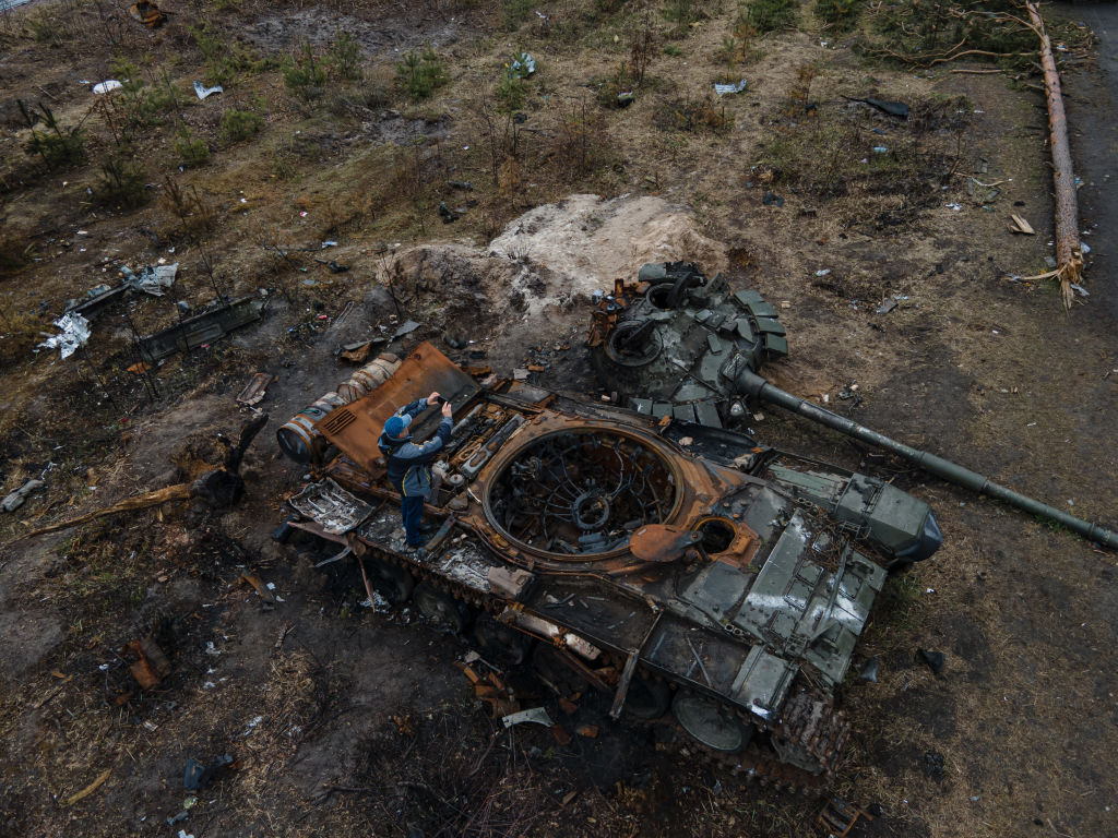 A photo of a man dressed in blue standing atop the shell of a destroyed Russian tank, taking a photo with his phone.