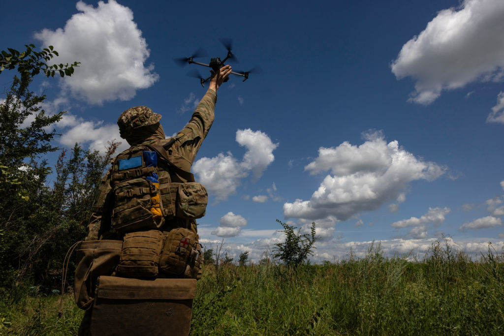 a photo of a man in full camouflage holding a quadcopter drone in a grassy field