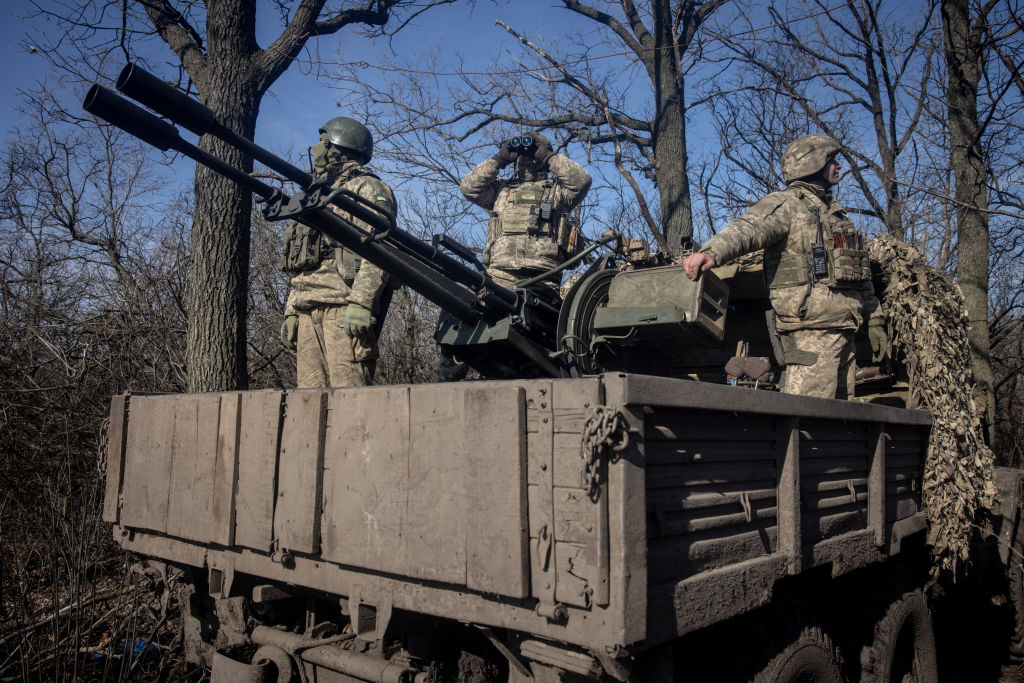 A photo of three people in military uniforms standing on the back of a truck next to a large anti-aircraft gun. One is looking up at the sky through binoculars.