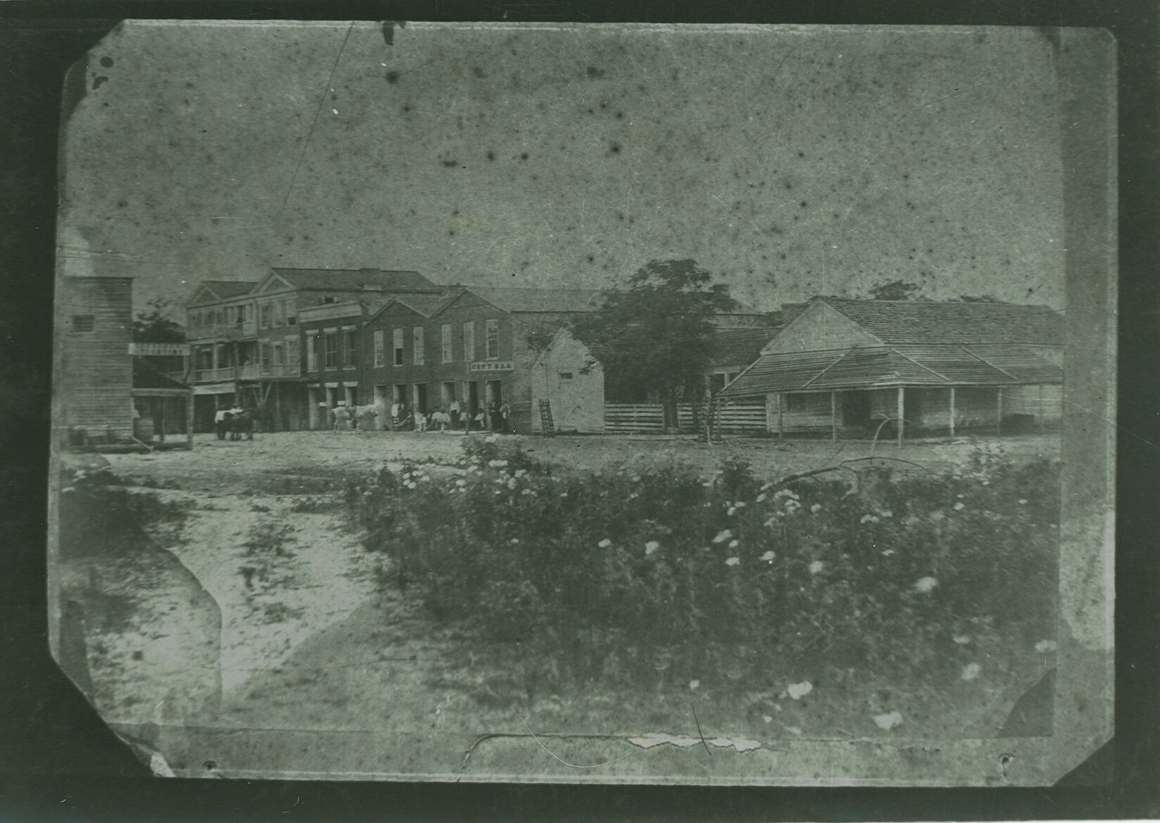 a black and white photo on a solid black background shows a row of buildings with people and animals in front of them