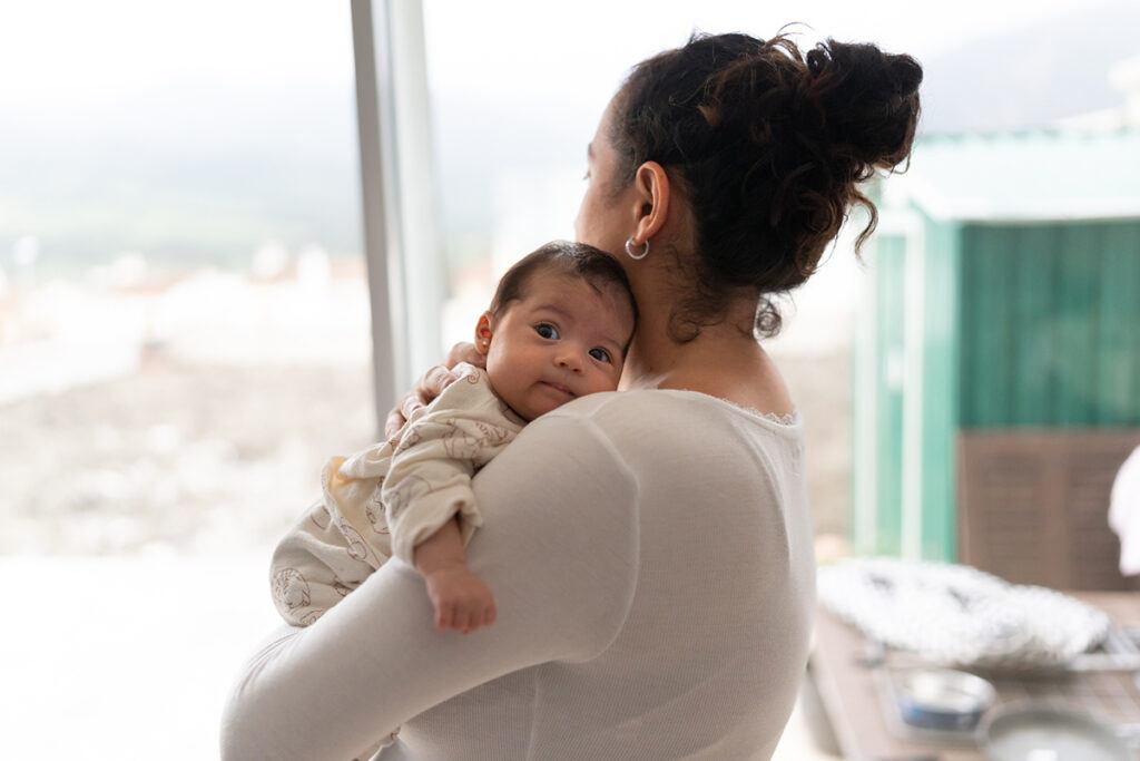 the mother is on her back looking out the window while holding the baby in her arms. The baby is looking at the camera.