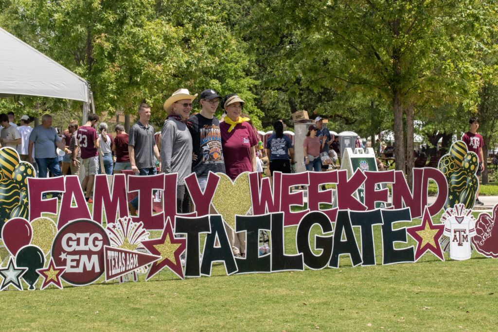 A family being photographed in Aggie Park behind a sign that says Family Weekend Tailgate.